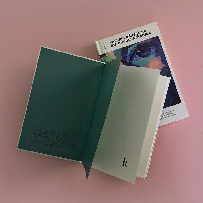 Foto von »Die Unvollständige«, ein Buch aufgeschlagen mit Klappentext im Vordergrund, Cover im Hintergrund.