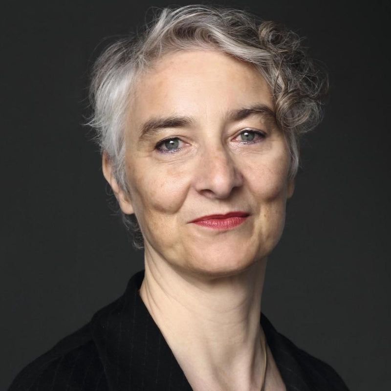 Porträt von Miriam Mandelkow. Sie trägt kurze graue Haare. Eine Locke hängt über dem rechten Auge. Miriam lächelt in die Kamera. Sie trägt ein schwarzes Oberteil mit Kragen. Eine Halskette ist im Ansatz sichtbar. Der Hintergrund ist dunkelgrau.