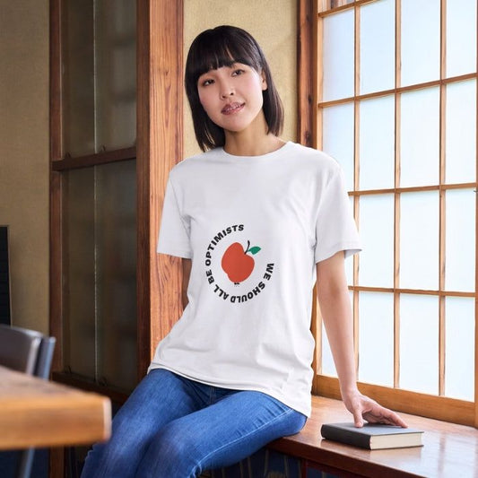 Nachhaltiges Feel good-T-Shirt We should all be optimists aus 100% Bio-Baumwolle mit rundem Print und Pfirsich-Motiv, getragen von junger Frau im Innenraum.