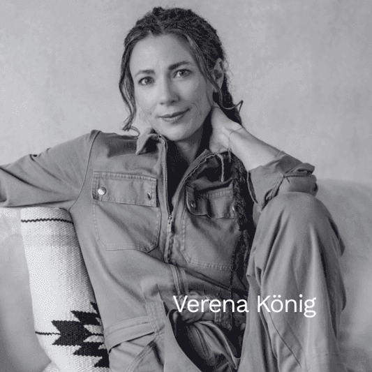 Schwarz-weiß Porträt der Autorin Verena König. Sie sitzt entspannt auf einem Sofa, trägt einen Overall und blickt mit einem sanften Lächeln direkt in die Kamera.