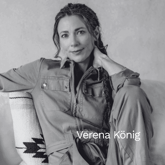 Schwarz-weiß Porträt der Autorin Verena König. Sie sitzt entspannt auf einem Sofa, trägt einen Overall und blickt mit einem sanften Lächeln direkt in die Kamera.