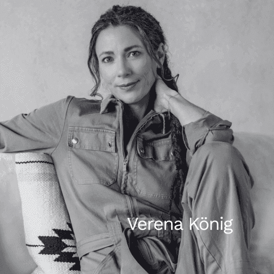 Schwarz-weiß Porträt der Autorin Verena König. Sie sitzt entspannt auf einem Sofa, trägt einen Overall und blickt mit einem sanften Lächeln direkt in die Kamera.