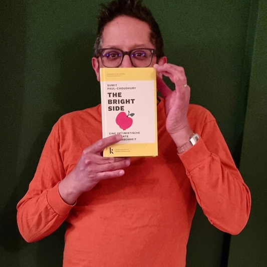 Der Autor Sumit Paul-Choudhury hält das Buch 'The Bright Side' vor grünem Hintergrund hoch, thematisiert Optimismus und menschlichen Fortschritt.