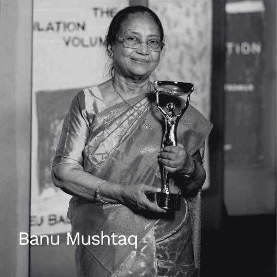 Schwarz-weiß Porträt der Autorin Banu Mushtaq. Sie trägt einen Sari und eine Brille, lächelt und hält eine Statuette (einen Buchpreis) in den Händen.