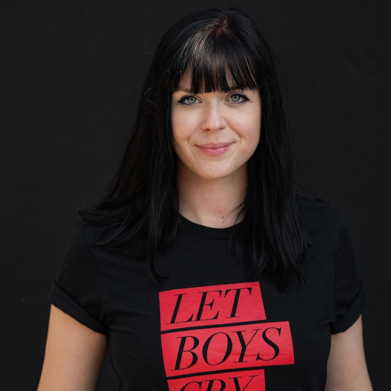 Porträt der Autorin Mareike Fallwickl. Sie lächelt in die Kamera. Schwarze Haare, schwarzes T-Shirt mit der Aufschrift "Let Boys Cry".