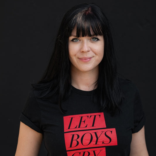 Porträt der Autorin Mareike Fallwickl. Sie lächelt in die Kamera. Schwarze Haare, schwarzes T-Shirt mit der Aufschrift "Let Boys Cry".