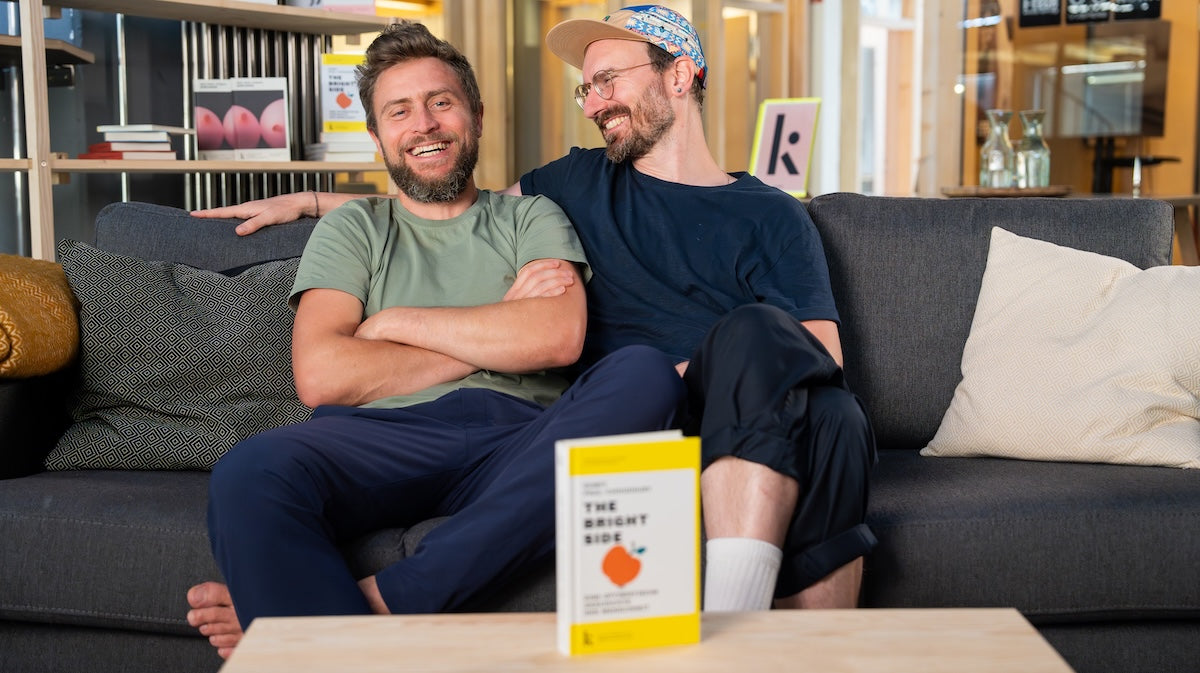 Kjona Gründer Lars (rechts) und Flo (links) sitzen fröhlich auf einer Couch und im Vordergrund steht das neue Buch über Optimismus "The Bright Side".