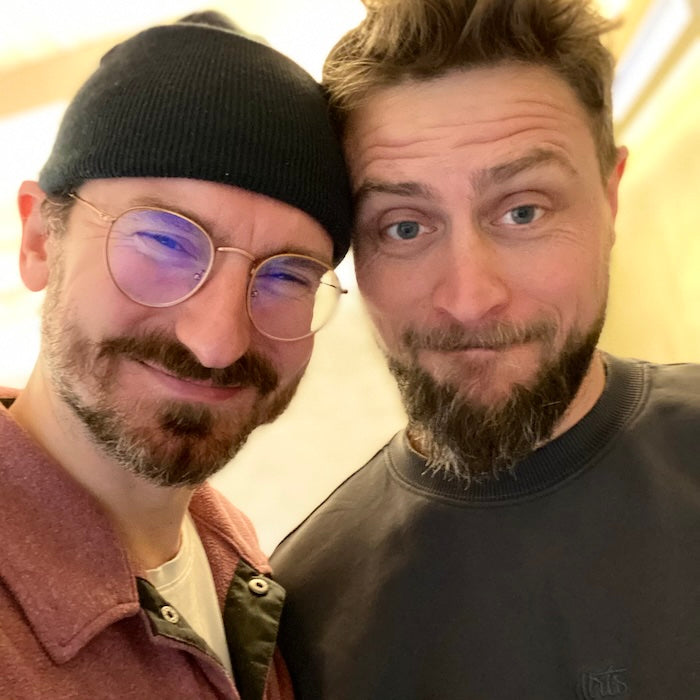 Lars (links) mit Mütze und Brille. Kurze Haare und Bart. Flo (rechts) mit Strubbelhaaren und Bart. Selfie im Workshop Raum des Impact Hubs München.