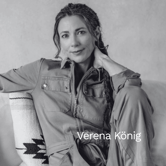 Schwarz-weiß Porträt der Autorin Verena König. Sie sitzt entspannt auf einem Sofa, trägt einen Overall und blickt mit einem sanften Lächeln direkt in die Kamera.